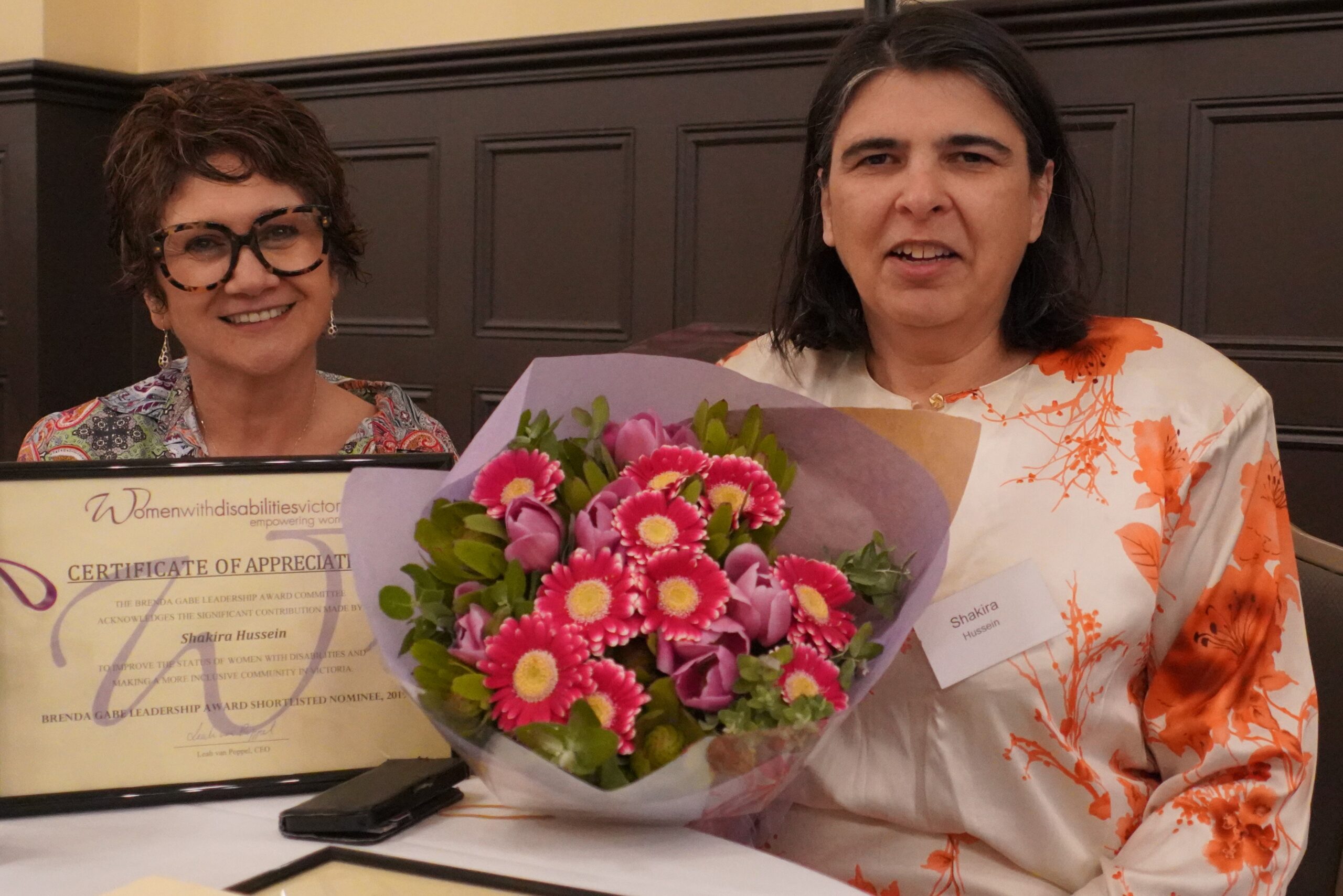 Two women sitting at a table. Shakira is on the left with a bunch of flowers in front of her. The lady sitting next to her is smiling and holding Shakira's certificate to the camera.