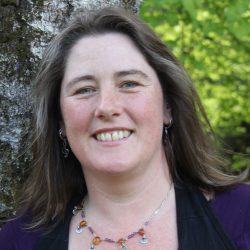 Close up photo of a white woman with wavy brown hair smiling at the camera. She is wearing a red necklace and the shoulders ofher black cardigan are visible in the frame.