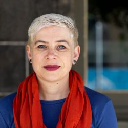 Headshot of a white woman with short blonde hair gazing at the camera with a closed mouth smile, wearing stud earrings, red lipstick, a red scarf and a blue top.