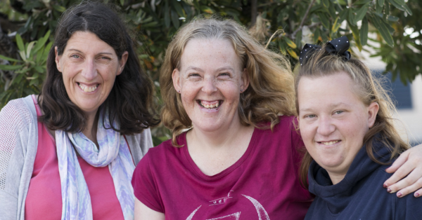 Three white women posing together, dressed casually, smiling for the camera, in front of a tree.