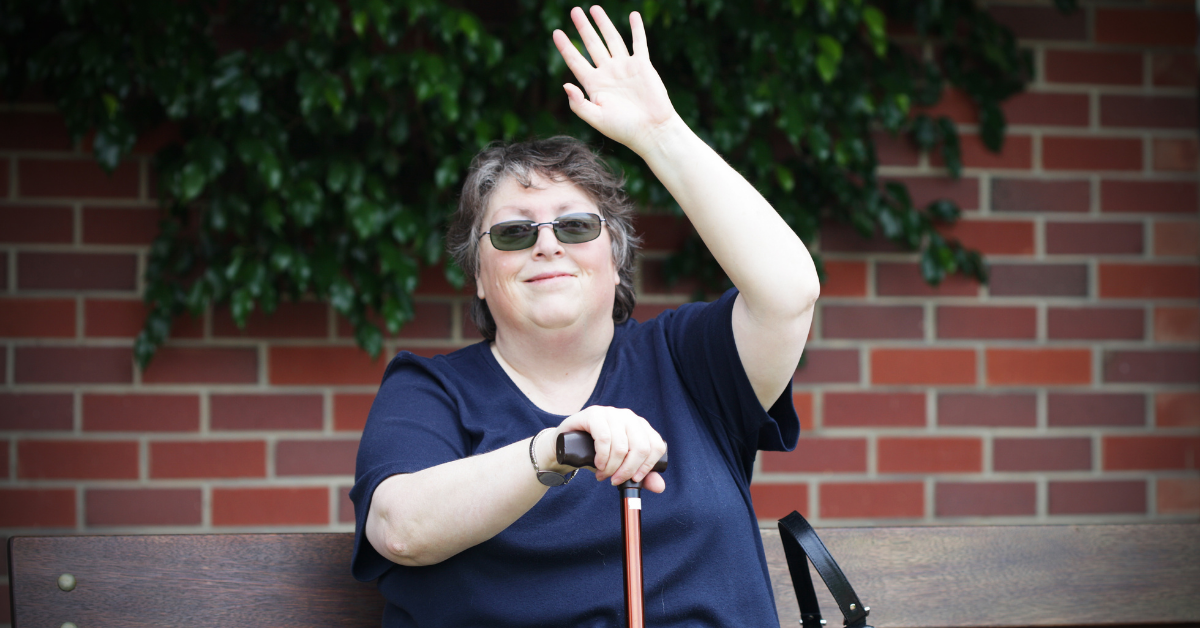 A white woman with short grey hair and sunglasses sits outise on a ench in front of a brick wall and tree. She holds a cane in her right hand. Her left hand is raised as though to ask a question.