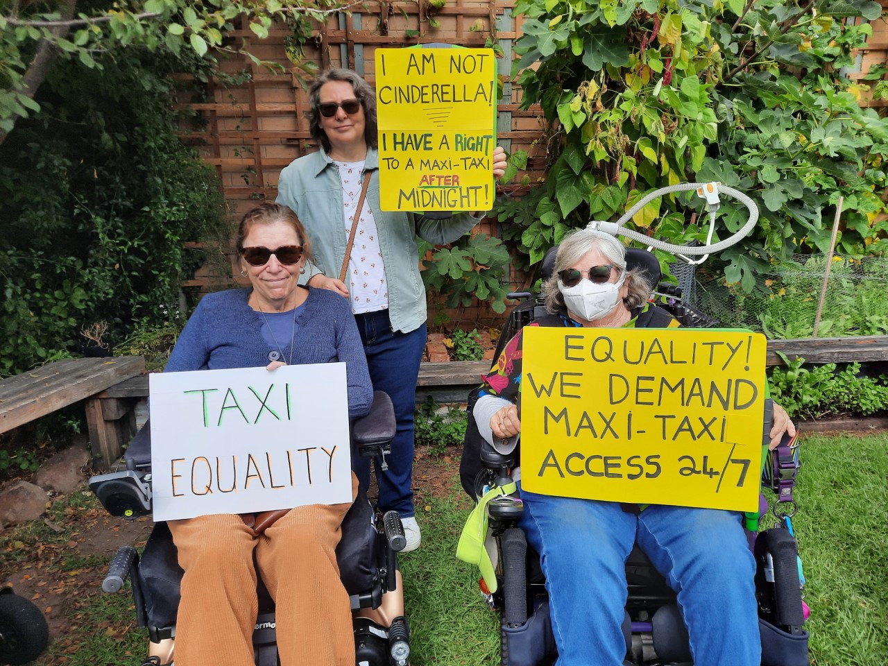 Three women in a backyard holding protest signs. Two of the women use motorised wheelchairs. One wears a mask. The signs say: "I am not Cinderella! I have a right to a maxi-taxi after midnight", "Taxi Equality" and "Equality! We demand maxi-taxi access 24/7".
