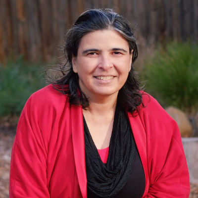 A smiling woman with dark hair and a red cardigan, seated outdoors in a wooded area.