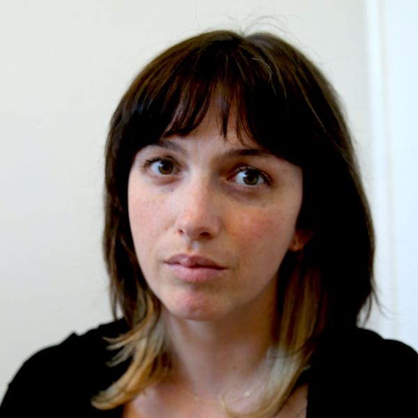 Close up photo of a woman looking seriously at the camera. She has shoulder length brown hair and a fringe.