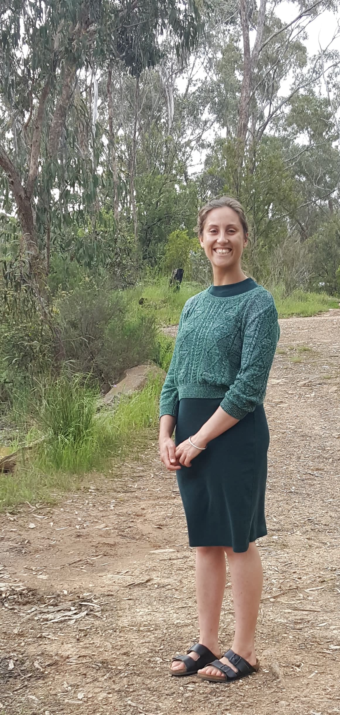 Marlena Raymond. A photo of a person standing outside with trees behind them. Person is smiling, standing and wearing green shirt and black skirt