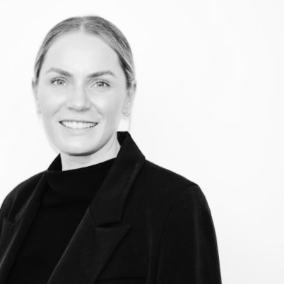 Black and white image of Rachel Arbon. She is wearing a black jumper, her blond hair is tied back and parted in the middle and she is looking at the camera smiling.