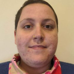 Photo of Nia, wearing a navy blue jumper with the collar of a colourful pink, tan and red shirt popping out. Nia's smiling at the camera.