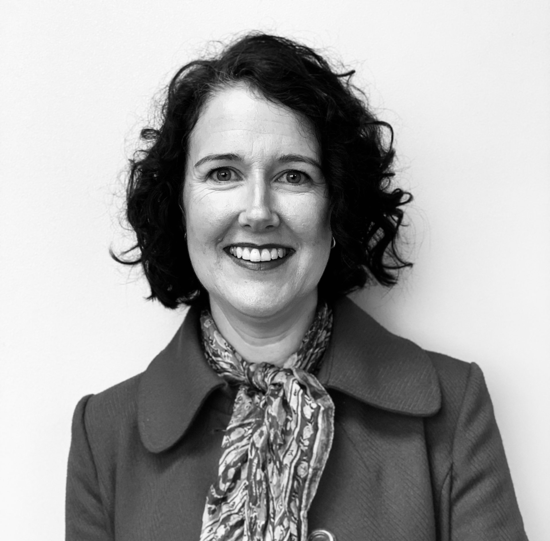 Photo of Melanie Guiney, in black and white. She is wearing a patterned scarf and a tailored coat. Her curly hair frames her face and she's smiling at the camera.