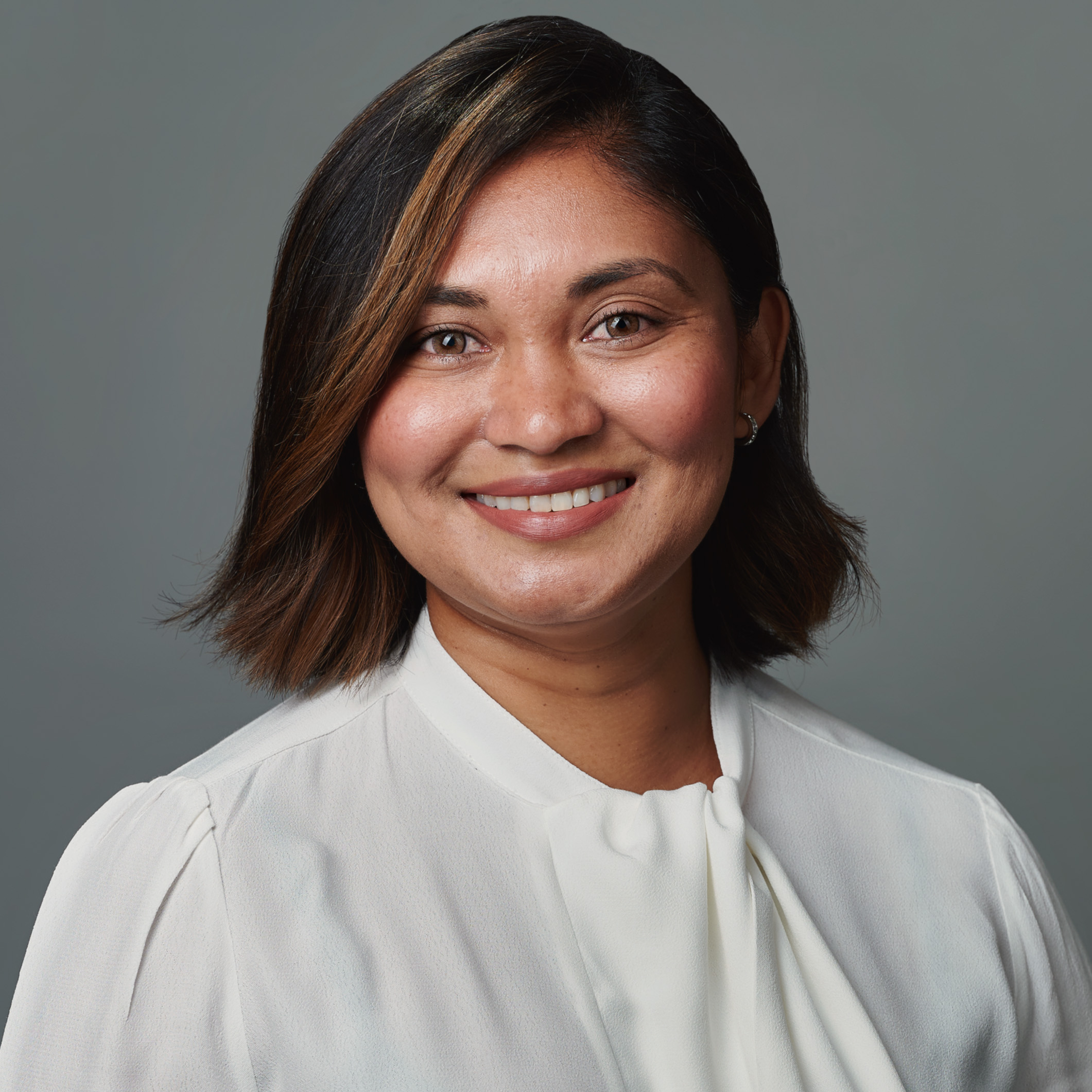 Photo with dark grey background. Woman of South-East Asian descent smiling, with shoulder length brown hair swept to one side and wearing a plain white shirt.