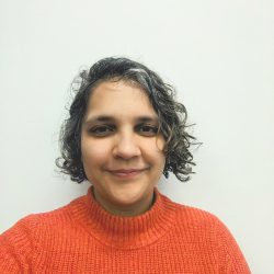 Saumya smiling at the camera, wearing a red/orange knitted jersey and curly hair.